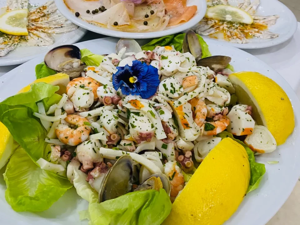 Antipasti di mare freschi al Ristorante Pizzeria Il Piacere di Modena: insalata di mare con polpo, gamberi, seppie e vongole, decorata con fiori ed erbe aromatiche, servita con spicchi di limone