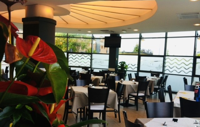 Interno del Ristorante Pizzeria Il Piacere a Modena, ambiente moderno con tavoli apparecchiati, fiori rossi decorativi e ampie vetrate. Specialità della casa: piatti a base di pesce